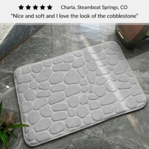 Light gray bath mat with cobblestone pattern placed on bathroom floor with customer review praising softness and design