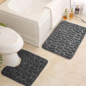 Dark gray pebble bath mat set featuring standard and contour shapes placed around white tub and toilet on beige tile floor