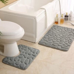 Light gray pebble bath mat set featuring standard and contour shapes placed around white tub and toilet on beige tile floor