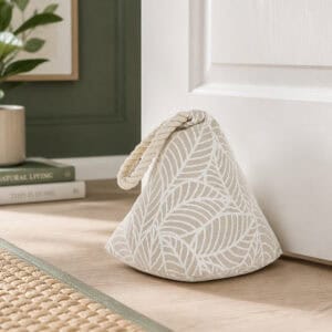 White leaf print cone door stopper propping open a white paneled door, hardwood floor, framed art, plant, and books in the background.
