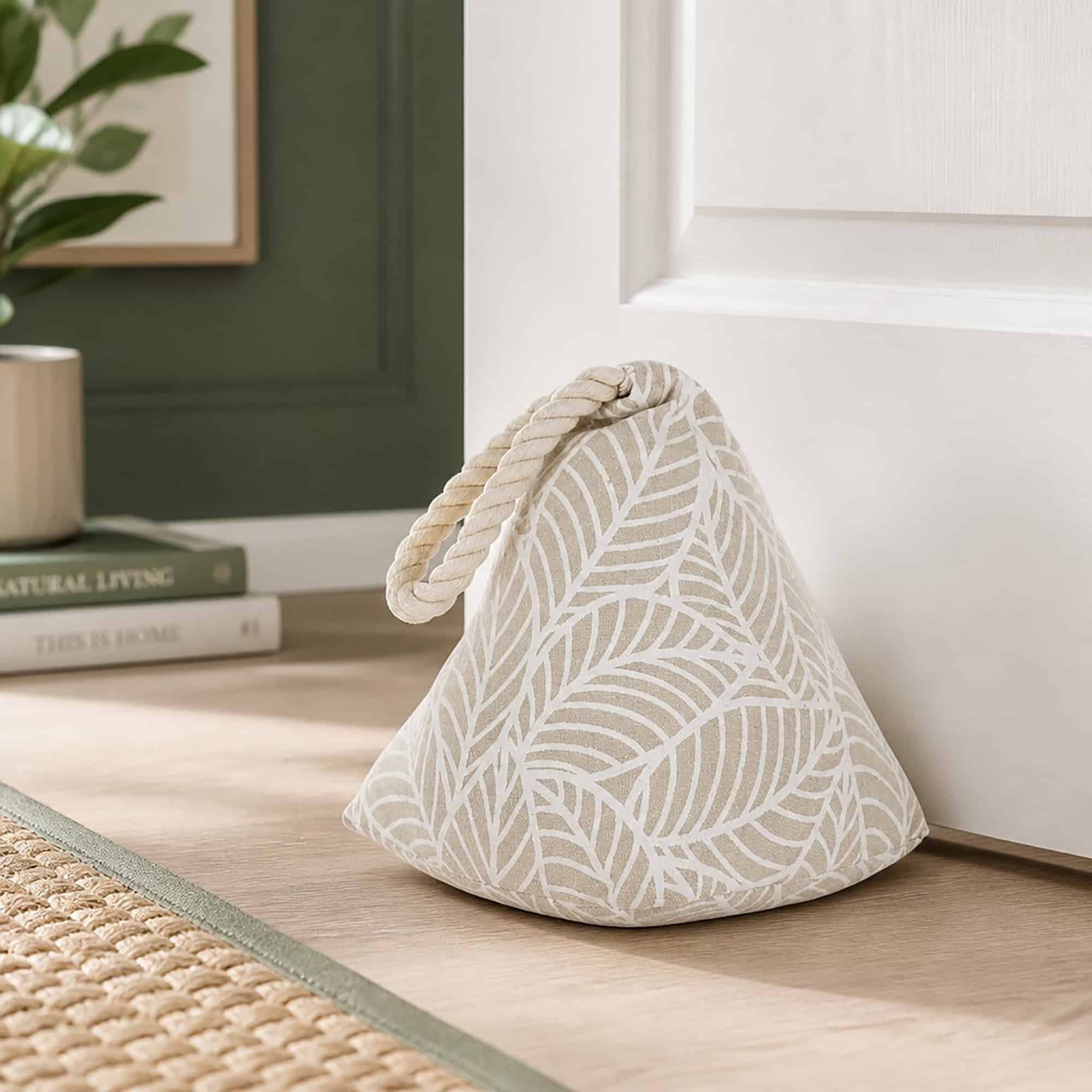 White leaf print cone door stopper propping open a white paneled door, hardwood floor, framed art, plant, and books in the background.