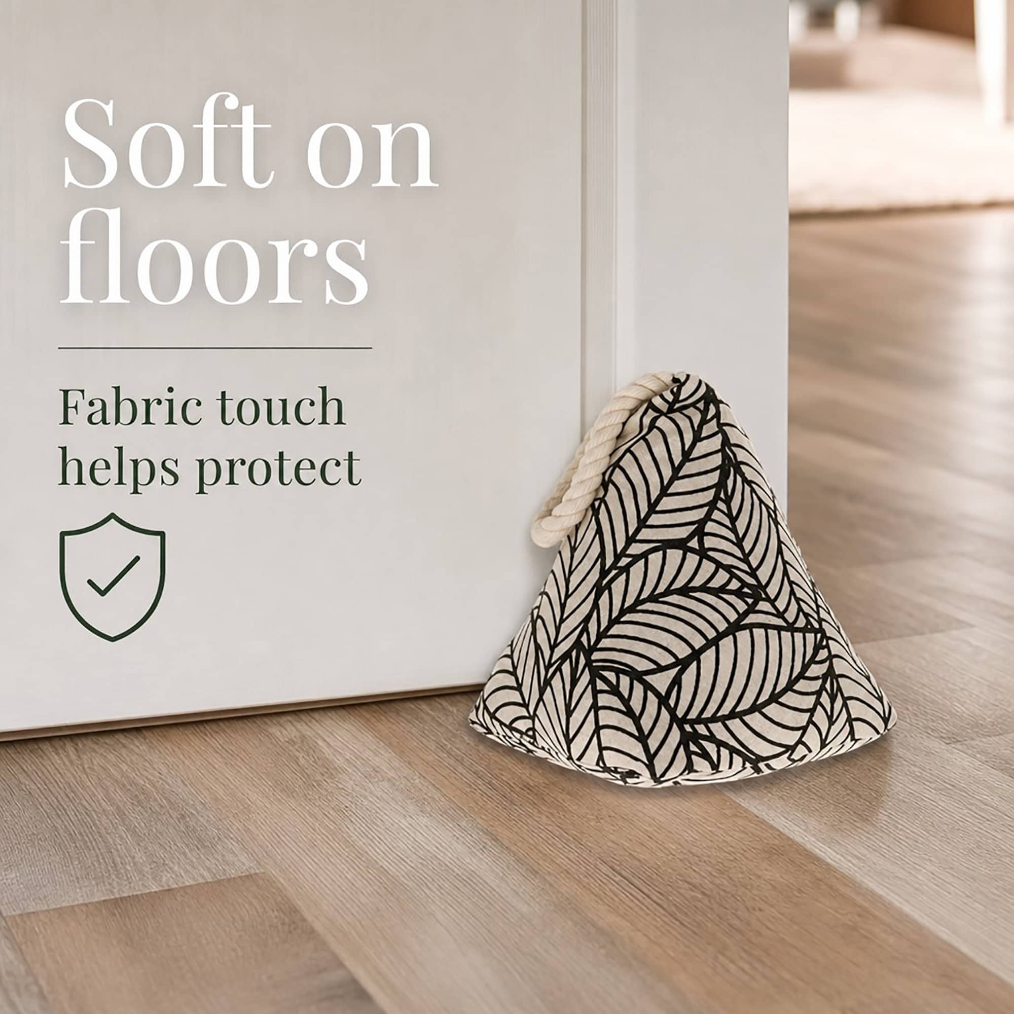 Black leaf print cone door stopper propping open a white door on hardwood floor, with a shield icon. On-image text: "Soft on floors," "Fabric touch helps protect."