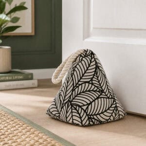 Black leaf print cone door stopper propping open a white paneled door, hardwood floor, framed art, plant, and books in the background.