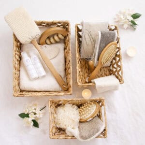 Overhead view of three water hyacinth baskets filled with spa and bath essentials including brushes, loofahs, and skincare products, styled with candles and white flowers.