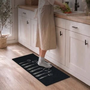 Person in a beige dress and white socks standing on a black cutlery runner rug while prepping food at a bright kitchen sink.