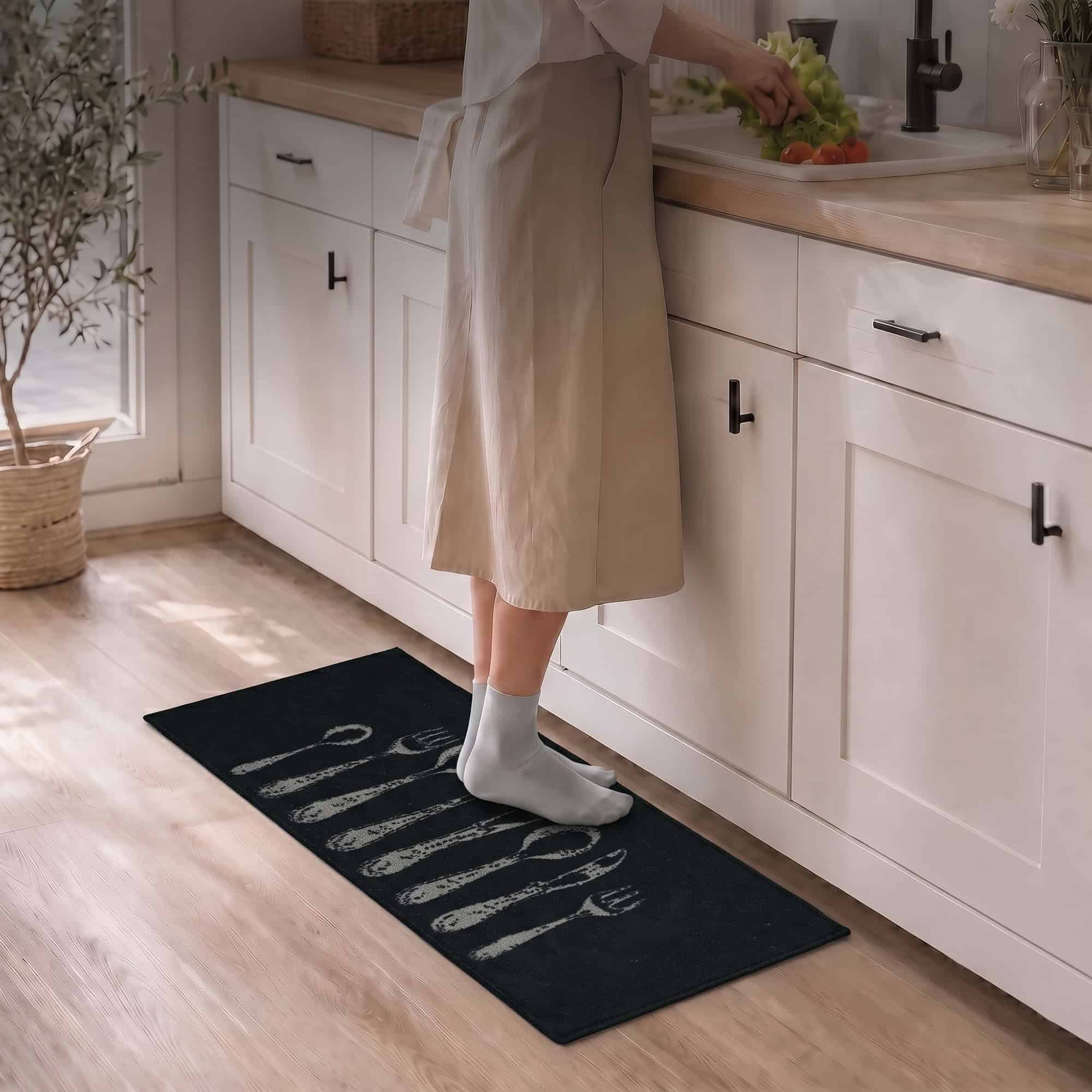 Person in a beige dress and white socks standing on a black cutlery runner rug while prepping food at a bright kitchen sink.