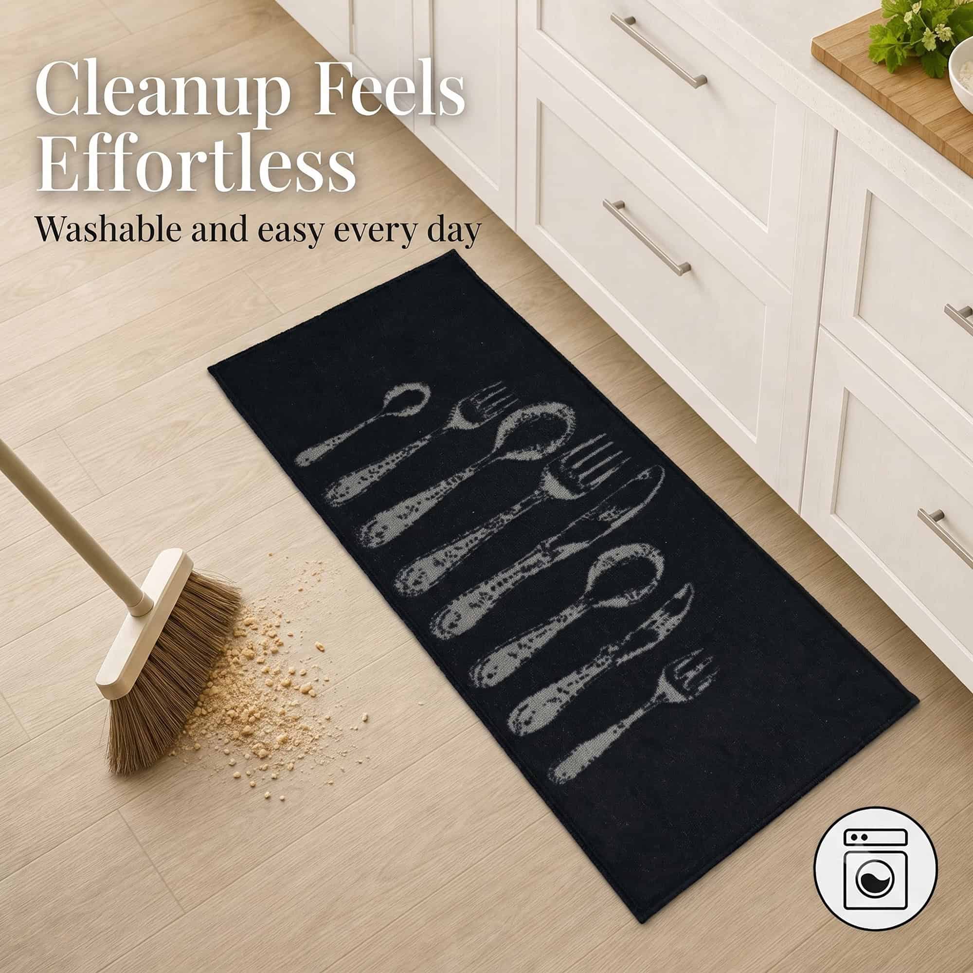 Black kitchen runner on a wood floor beside a broom sweeping crumbs, showing the washable surface that makes everyday cleanup feel effortless.