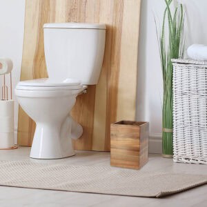 A wooden waste basket placed on the floor near a toilet in a bathroom.