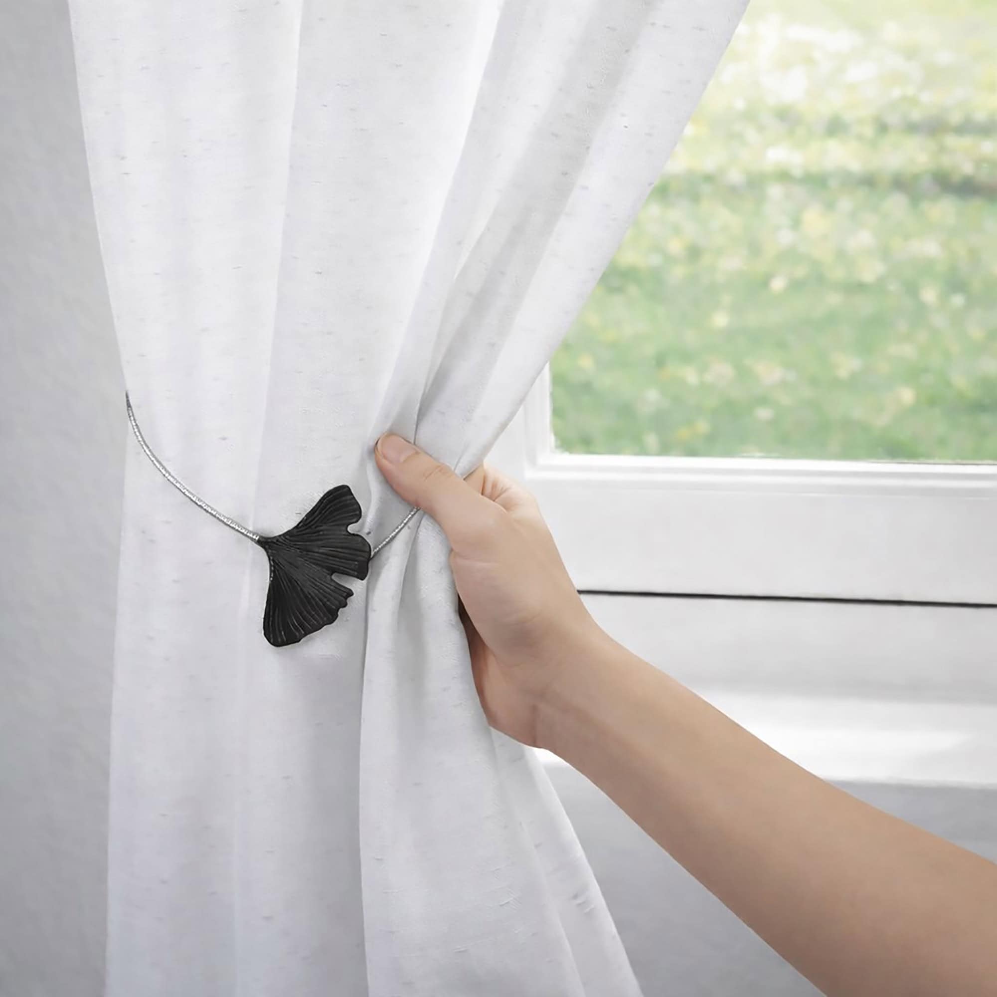 "Hand attaching black ginkgo leaf magnetic tieback to white curtain at window"