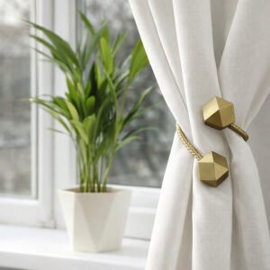 Gold geometric tieback bundling a white curtain near a sunlit window with indoor plant