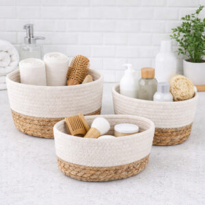 Three white cotton and seagrass baskets filled with rolled towels, hairbrushes, skincare bottles, and a loofah on a bathroom countertop.