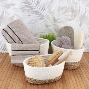 Three white cotton and seagrass baskets filled with towels, bath brushes, loofahs, and spa accessories on a bamboo surface.