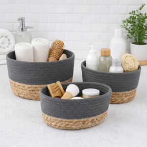 Three gray cotton and seagrass baskets filled with rolled towels, hairbrushes, skincare bottles, and a loofah on a bathroom countertop.