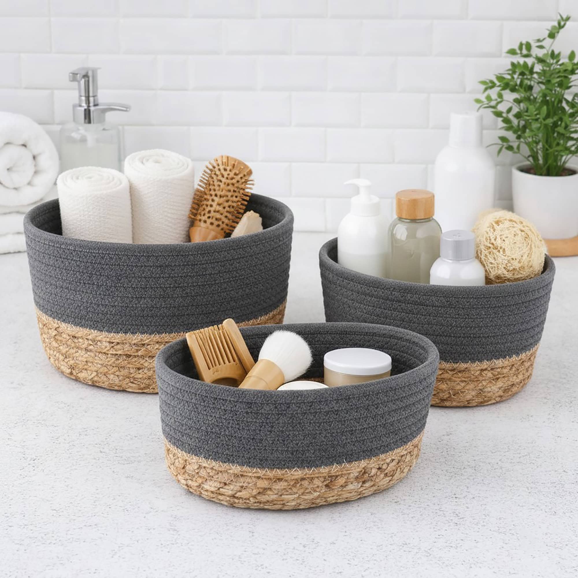 Three gray cotton and seagrass baskets filled with rolled towels, hairbrushes, skincare bottles, and a loofah on a bathroom countertop.