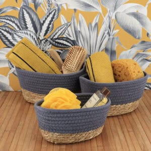 Three gray cotton and seagrass baskets filled with yellow towels, bath brushes, a loofah, and shower gel on a bamboo surface with tropical wallpaper backdrop.