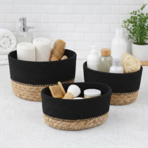 Three black cotton and seagrass baskets filled with rolled towels, hairbrushes, skincare bottles, and a loofah on a bathroom countertop.