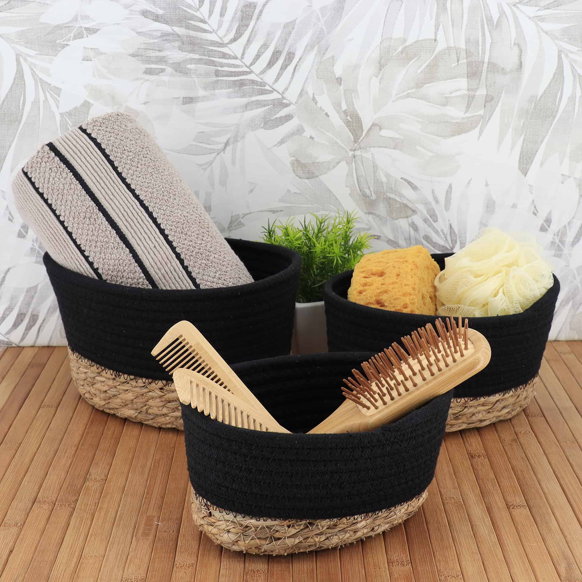 Three black cotton and seagrass baskets filled with towels, bath sponges, loofahs, and wooden hairbrushes on a bamboo surface with tropical wallpaper backdrop.