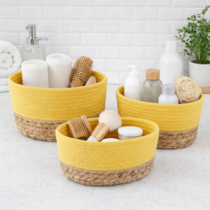 Three yellow cotton and seagrass baskets filled with rolled towels, hairbrushes, skincare bottles, and a loofah on a bathroom countertop.