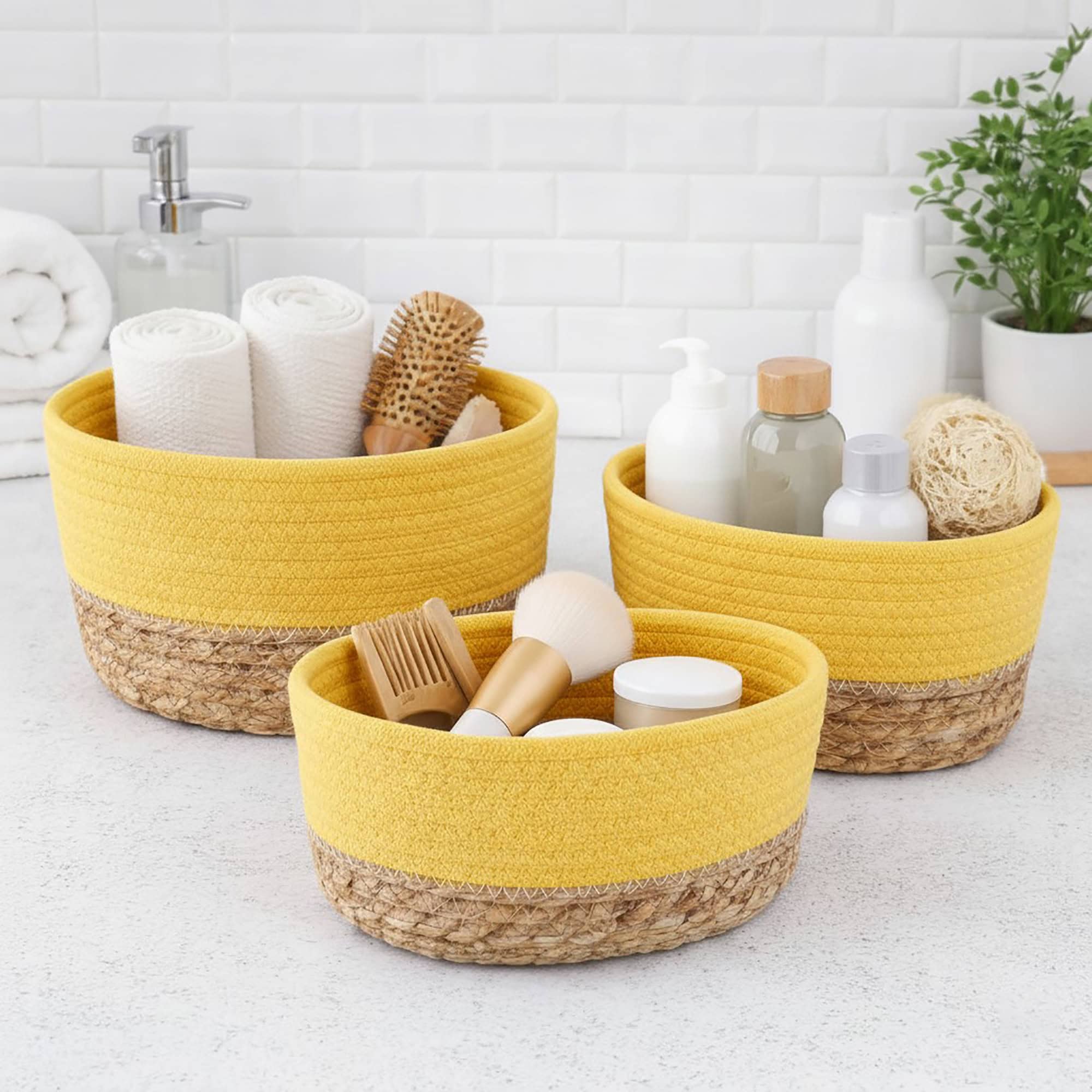 Three yellow cotton and seagrass baskets filled with rolled towels, hairbrushes, skincare bottles, and a loofah on a bathroom countertop.