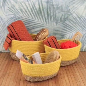 Three yellow cotton and seagrass baskets filled with orange towels, bath brushes, a loofah, and a pumice stone on a bamboo surface with tropical wallpaper backdrop.