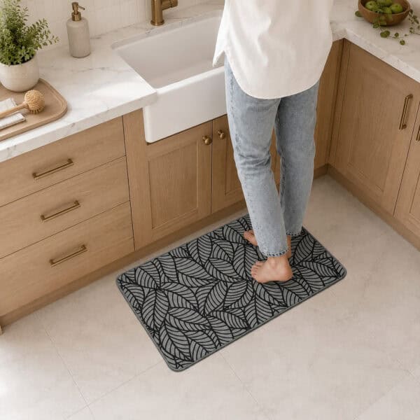 Person standing barefoot on a grey leaf-print kitchen mat in front of a farmhouse sink in a bright wood-cabinet kitchen.