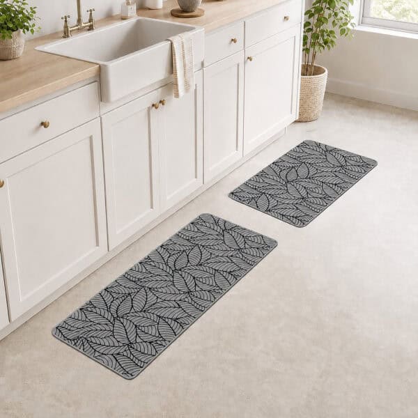 Grey leaf-print kitchen mat and runner placed together in front of a white farmhouse sink in a bright, plant-filled kitchen.