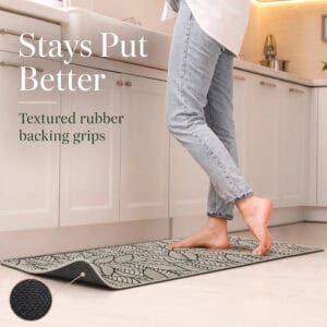 Person stepping onto a beige leaf-print kitchen mat, the lifted corner revealing the textured rubber backing that grips the floor.