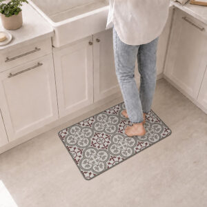 A person stands barefoot on the kitchen mat in front of a white farmhouse sink, the red and gray tile pattern adding warmth to the space.