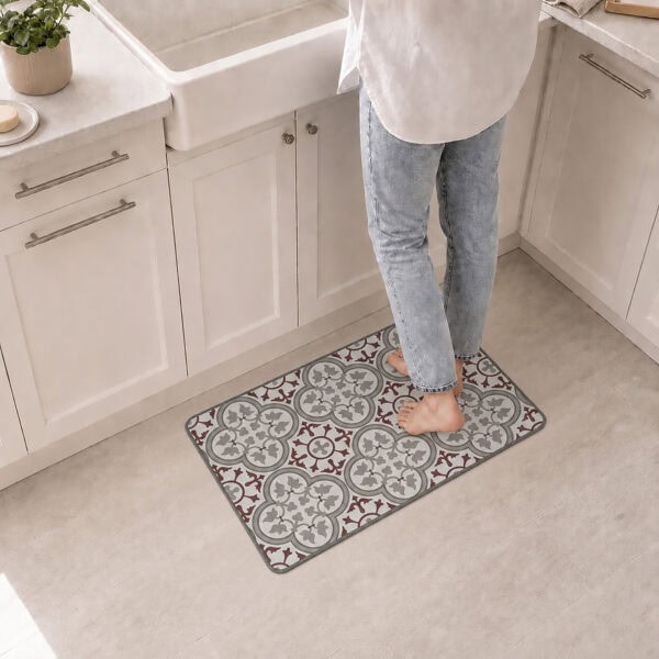 A person stands barefoot on the kitchen mat in front of a white farmhouse sink, the red and gray tile pattern adding warmth to the space.