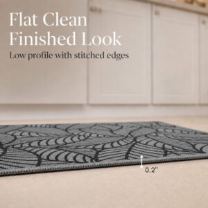 Side view of a grey leaf-print kitchen mat lying flat, showing its thin two-tenths of an inch profile with stitched edges.