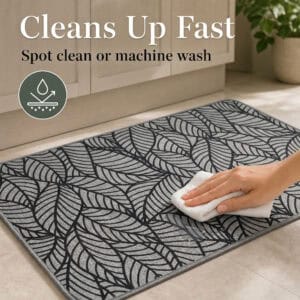 Hand wiping a grey leaf-print kitchen mat with a white cloth, showing it can be spot cleaned or machine washed.