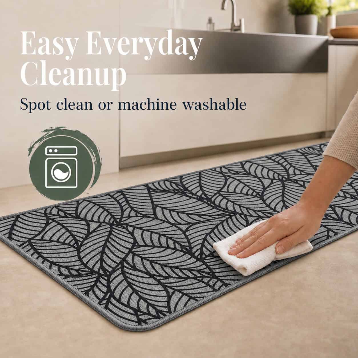 Hand wiping a grey leaf-print kitchen runner with a white cloth, showing it can be spot cleaned or machine washed.