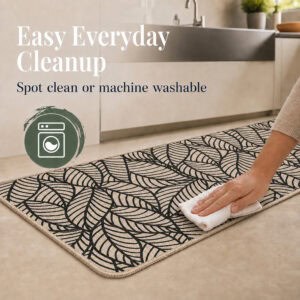 Hand wiping a beige leaf-print kitchen mat with a white cloth, showing the set can be spot cleaned or machine washed.