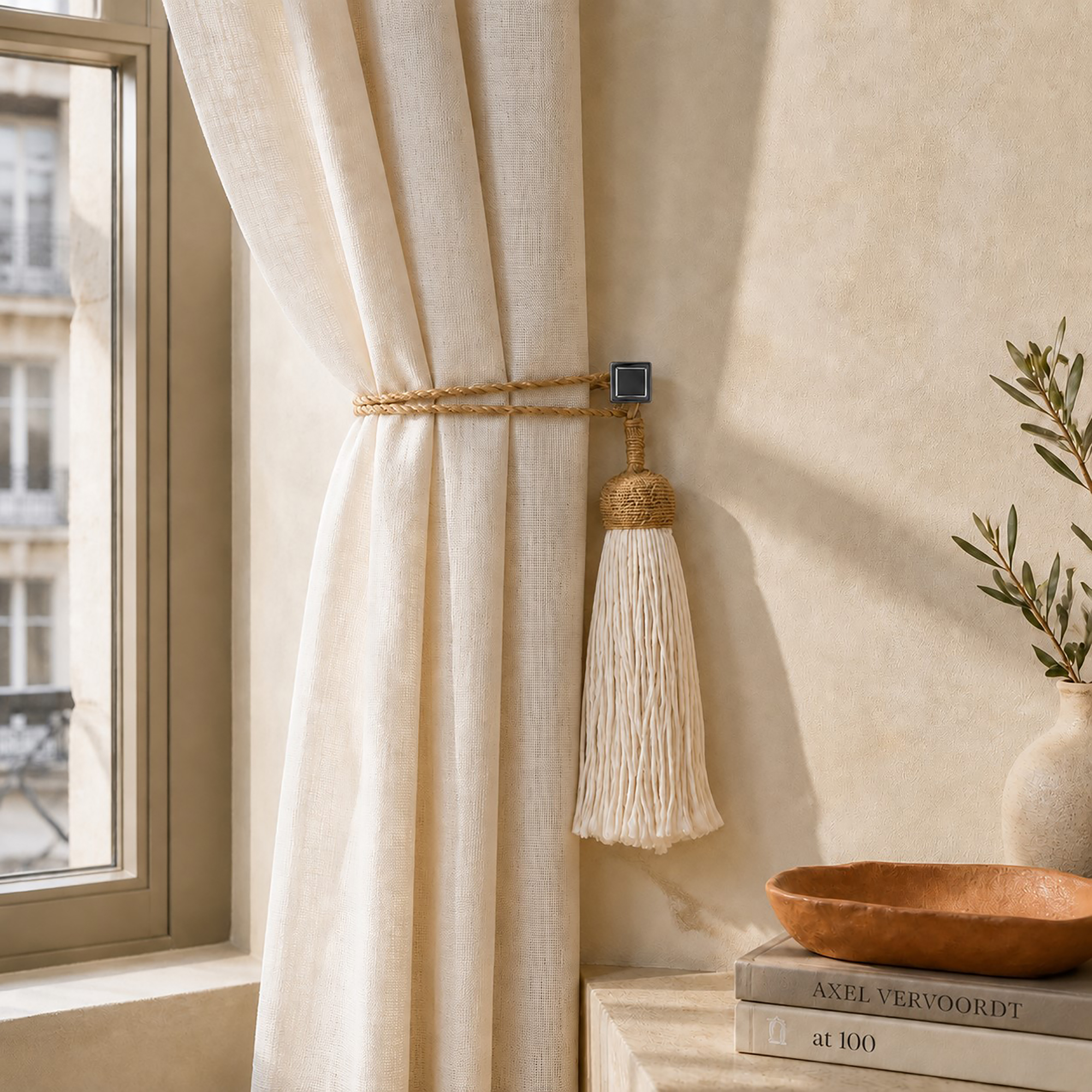 Graphite square tieback hook holding an ivory linen curtain with a gold rope and tassel in a sunlit room