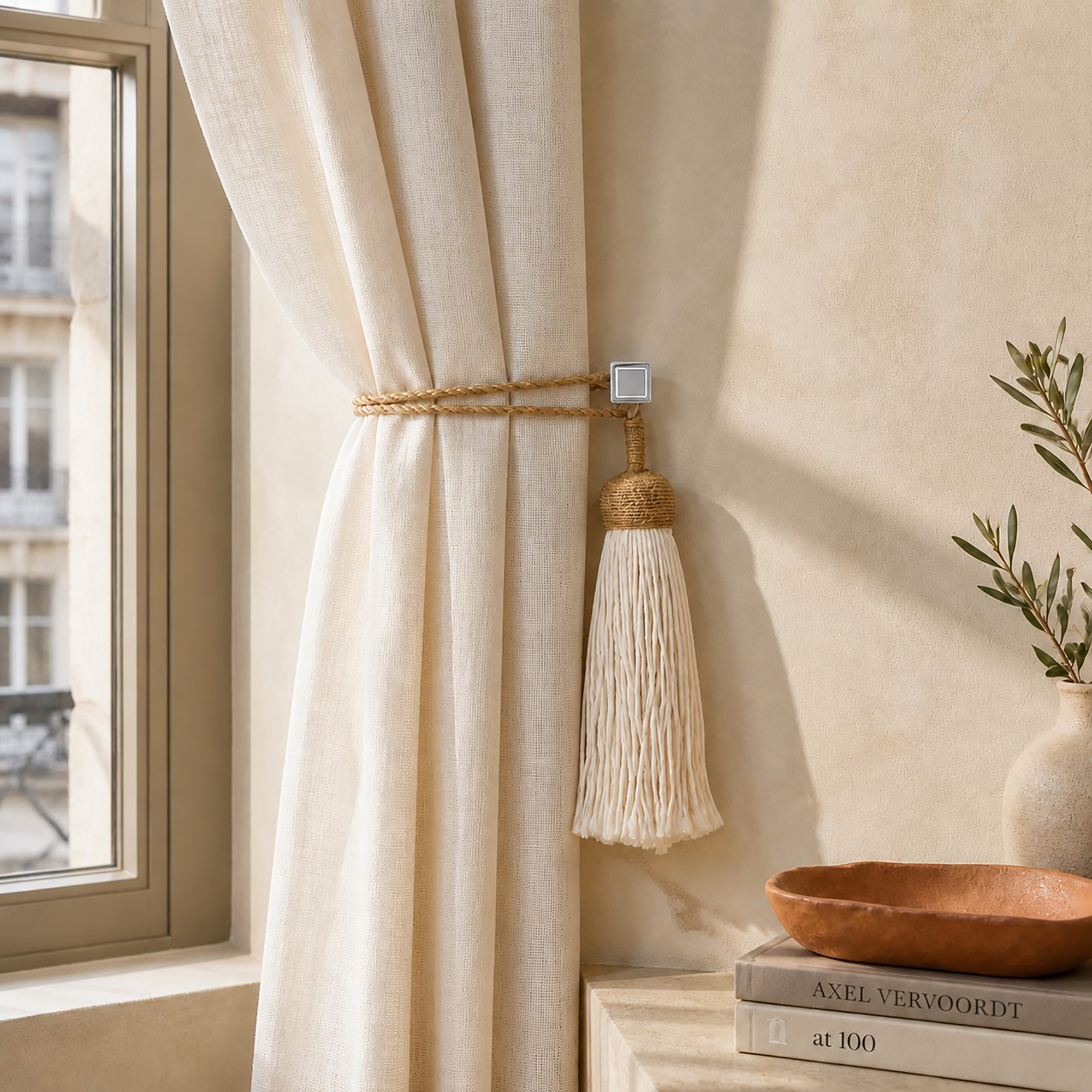 Chrome square tieback hook holding an ivory linen curtain with a gold rope and tassel in a sunlit room