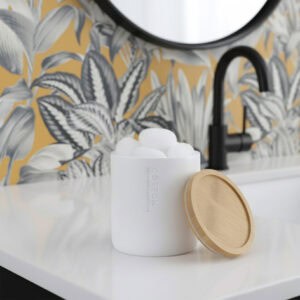 White matte cotton ball jar with bamboo lid on bathroom countertop, displayed against yellow and gray tropical leaf wallpaper