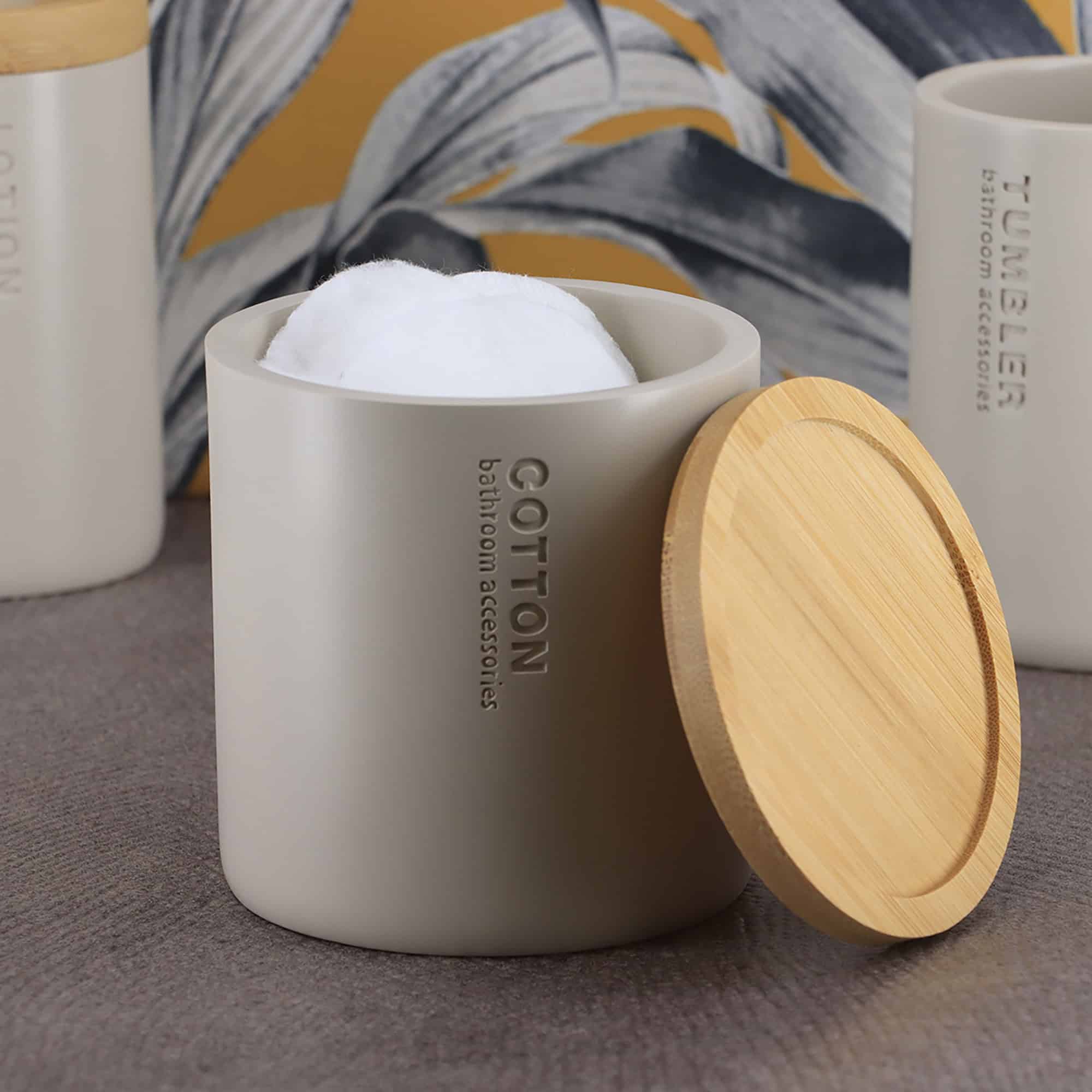 Open beige cotton jar with a bamboo lid, filled with cotton pads, styled with combs and rolled towels against a botanical wallpaper.