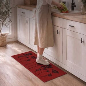 Woman in soft shoes standing on a red floral kitchen runner at the sink while rinsing fresh greens in a bright modern kitchen.