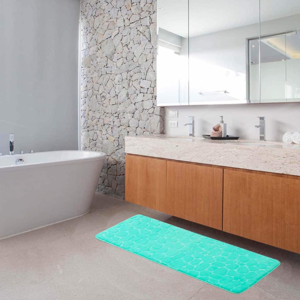 A plush aqua blue memory foam bath mat with a 3D pebble pattern lies on the floor of a modern bathroom, contrasting against a stone wall and sleek wooden vanity