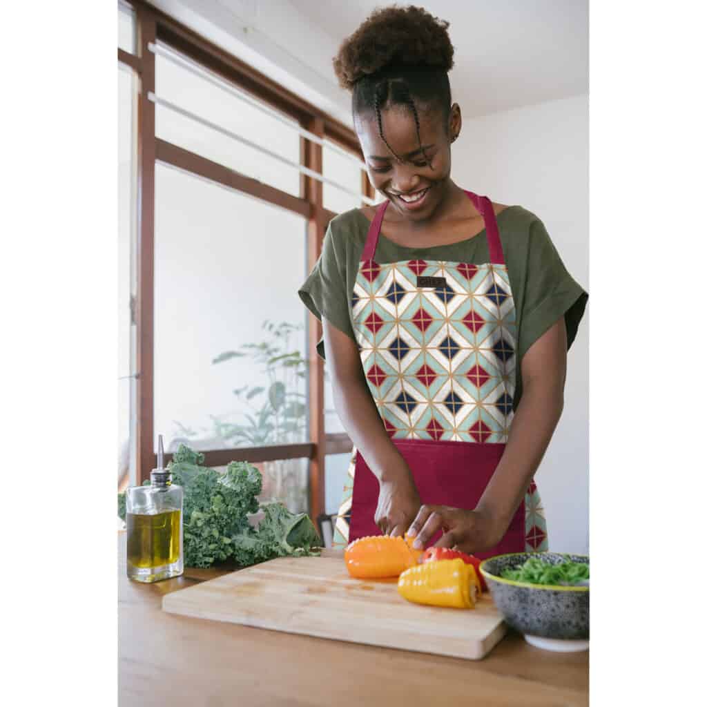  "A woman with a cheerful smile is chopping yellow bell peppers on a wooden cutting board, wearing a multicolored geometric apron, part of a 4-piece kitchen set, in a bright, plant-filled kitchen