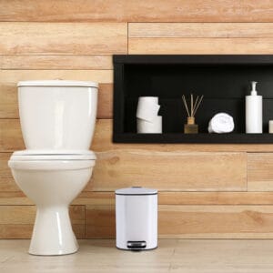 a white trash can next to toilet seat in bathroom environment