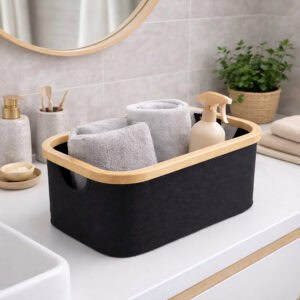 Black bamboo-rimmed storage basket holding rolled gray towels and a spray bottle on a bathroom countertop.