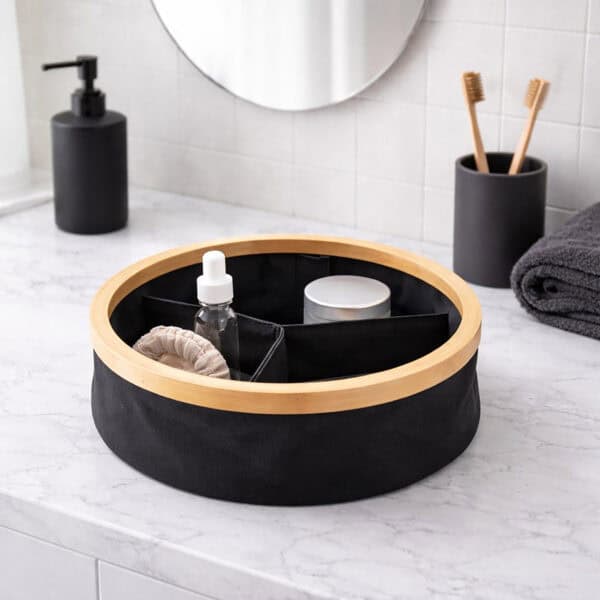Image 2: Black round bamboo-rimmed 3-compartment basket holding skincare products and a cotton scrunchie on a marble bathroom countertop.