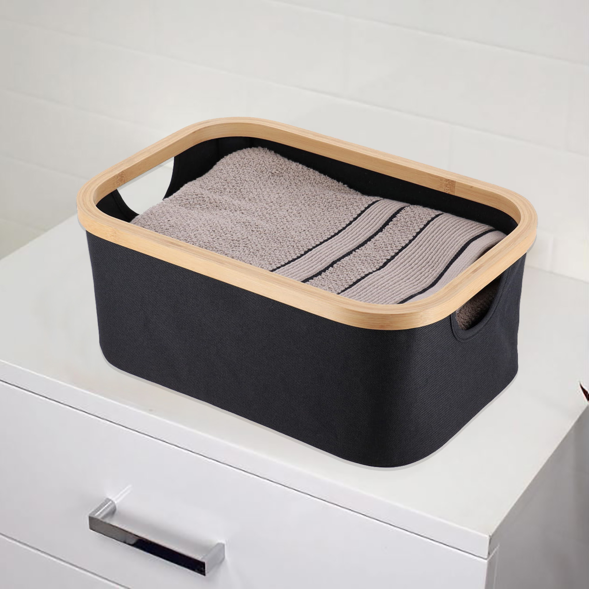 Black fabric storage basket with bamboo rim containing folded gray towels, placed on top of a white dresser.