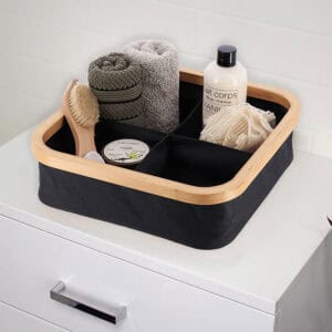 Black 4-compartment bamboo-rimmed basket holding bath accessories including rolled towels, a brush, and loofah on a white dresser.