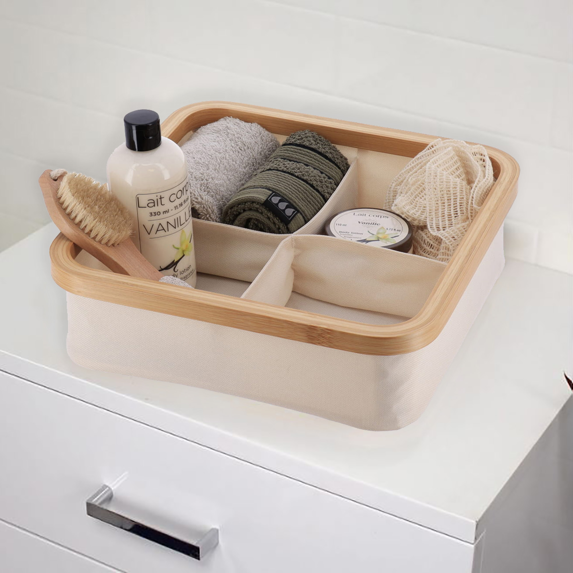 Ecru 4-compartment bamboo-rimmed basket holding bath accessories including body lotion, rolled towels, and a loofah on a white dresser.