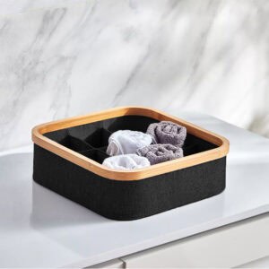 Image 7: Black bamboo-rimmed 9-compartment basket holding rolled white and gray towels, placed on a white surface against a marble wall.