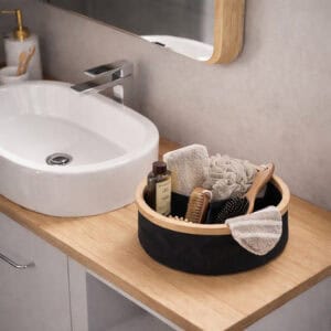 Image 7: Black round bamboo-rimmed basket holding bath accessories including brushes, a loofah, and shower gel on a wooden bathroom vanity beside a vessel sink.