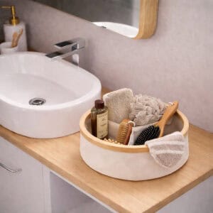 Image 1: Ecru round bamboo-rimmed basket holding bath accessories including brushes, a loofah, and shower gel on a wooden bathroom vanity beside a vessel sink.