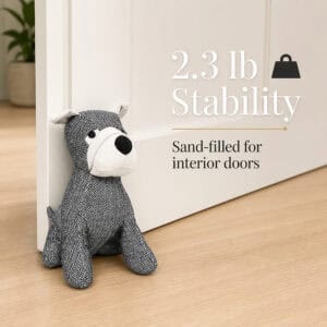 Charcoal dog door stopper propping open a white door on hardwood floor, potted plant behind, with a weight icon. On-image text: "2.3 lb Stability," "Sand-filled for interior doors."